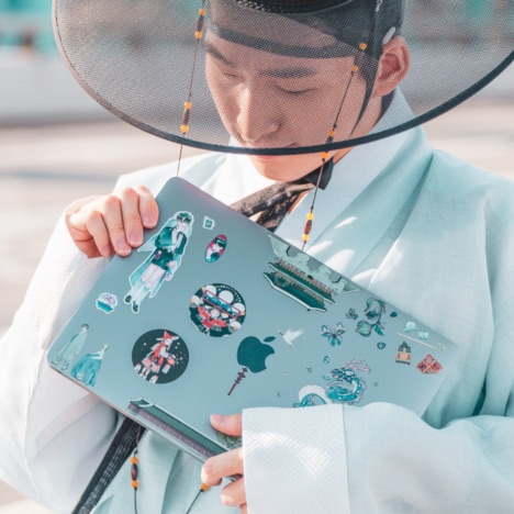 SuhJae wearing a sky-blue Hanbok (소창의, traditional Korean clothing) and a gat while holding a MacBook decorated with Korean-themed stickers.