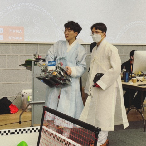 SuhJae in a blue Hanbok and teammate in a yellow Hanbok, holding a competition robot at the VEX Over Under Challenge.