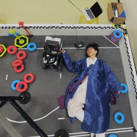SuhJae wearing a blue Hanbok (소창의), observing his robot during the VEX Robotics development phase.